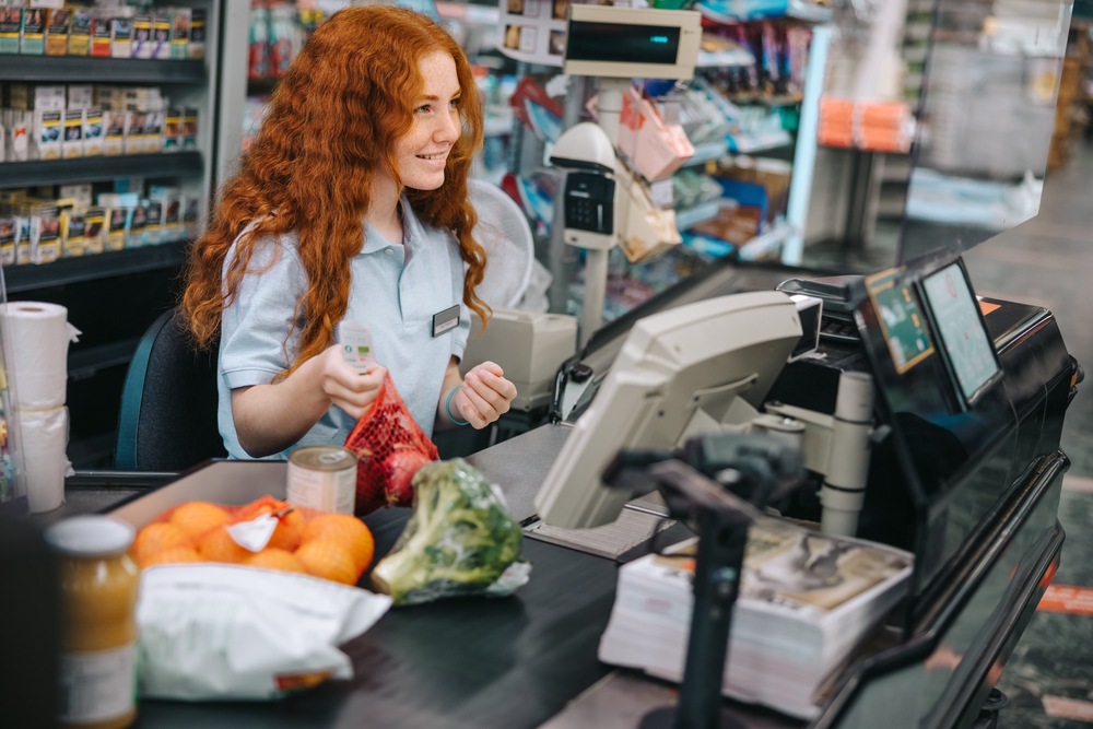 Young,Sales,Clerk,Sitting,By,Cash,Register,In,Supermarket,And